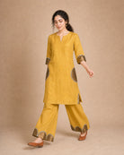 Front three-quarter view of the Arpita golden-mustard linen co-ord, with semicircle antique-shimmer and mirror-lace treatment visible on the sleeve and palazzo